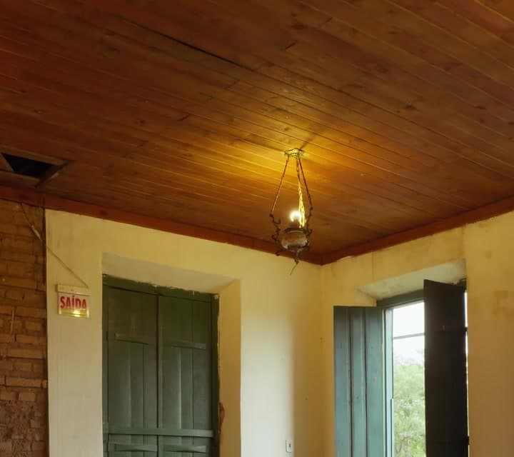 Sala do casarão com paredes amarelas, janelas verdes e lampião